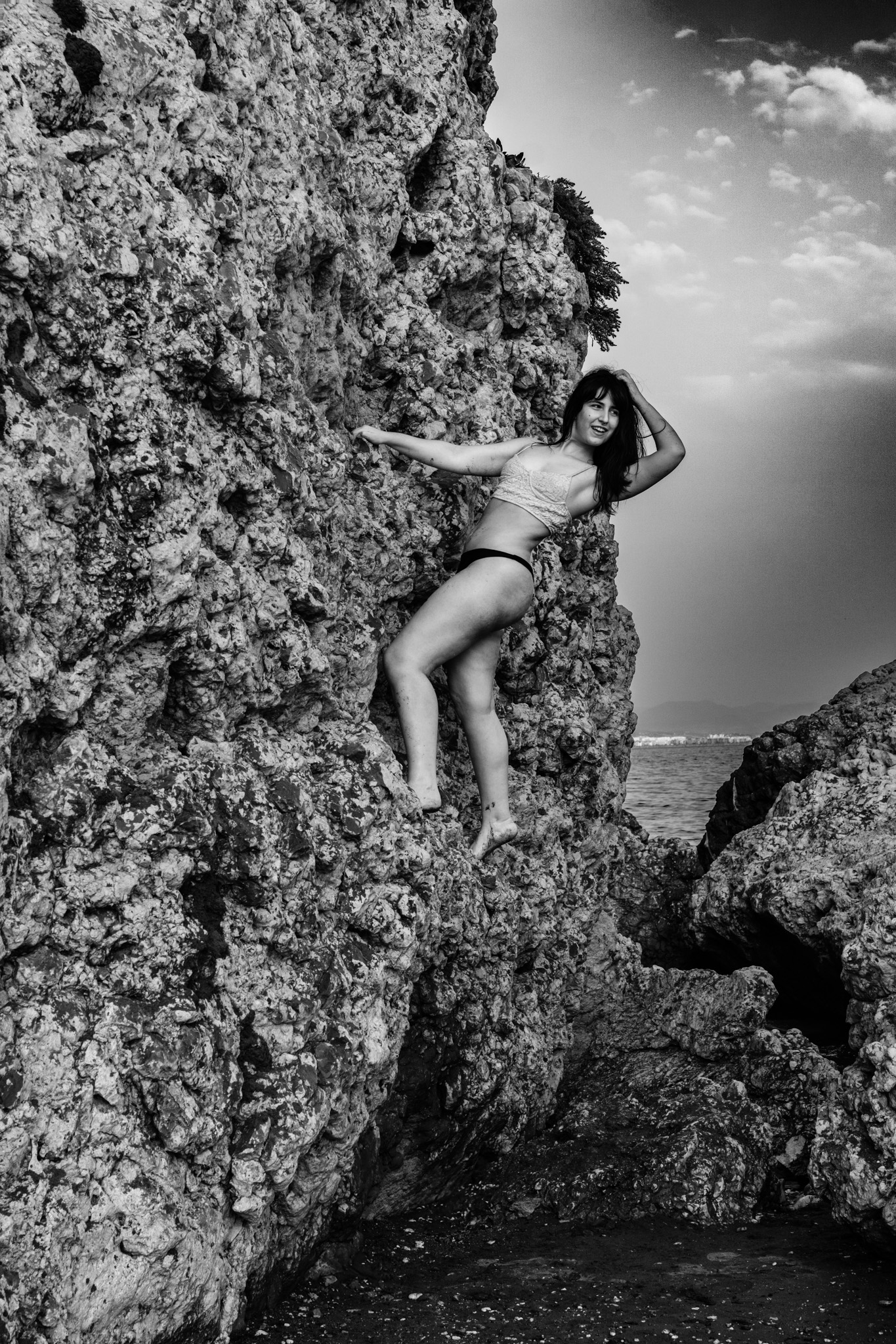 Woman scaling a craggy sea cliff between two rock faces, reaching upward, ocean and open sky behind her, black and white