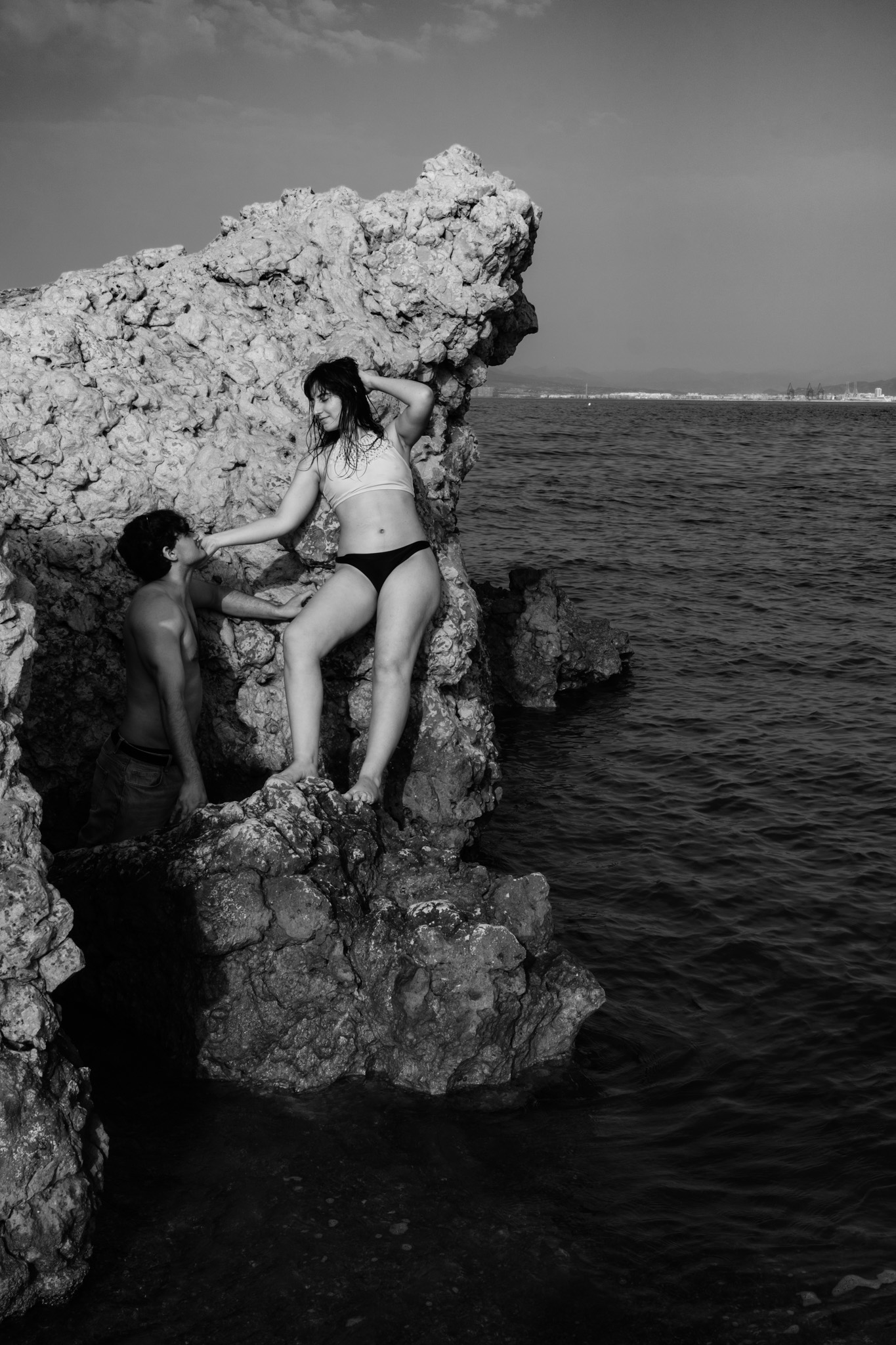 Woman poised on a sea cliff in a white crop top and black bikini bottoms, a man steadying her from the rocks below, black and white