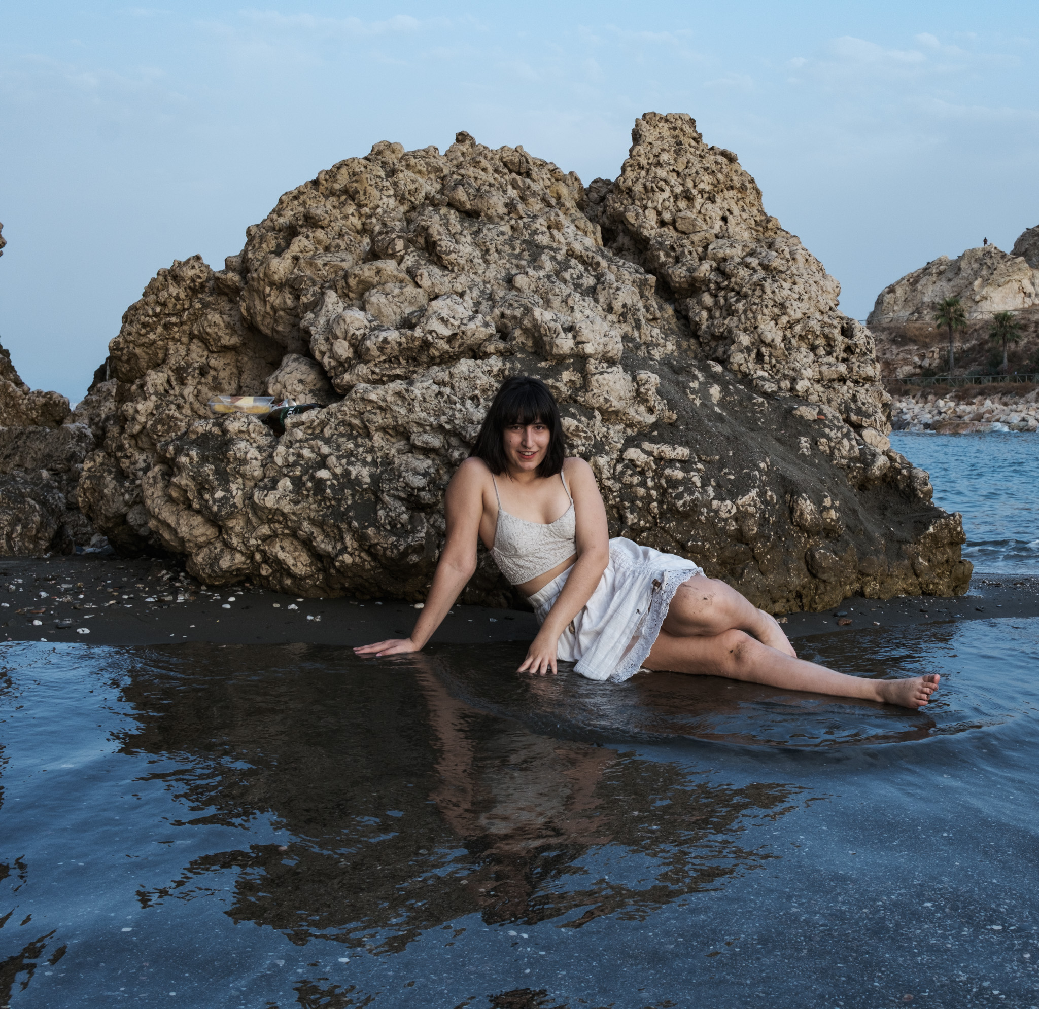 Woman seated in a shallow tidal pool between sea-worn rocks, wearing white lace, the still water reflecting the evening sky