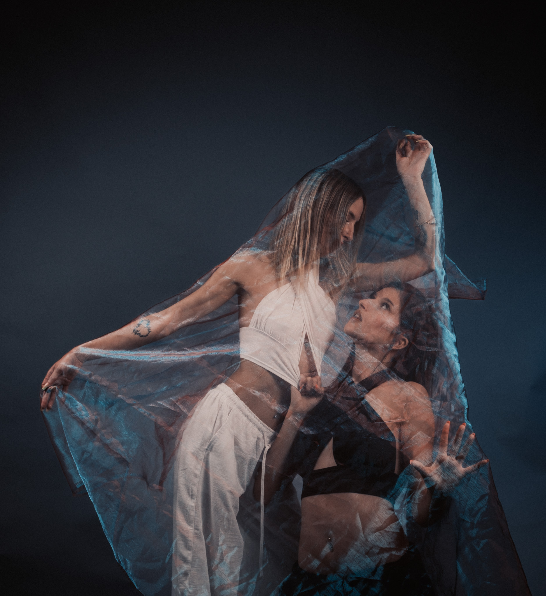 Two women in a moody dark studio lifting a large translucent blue veil overhead, their forms visible through the layered fabric