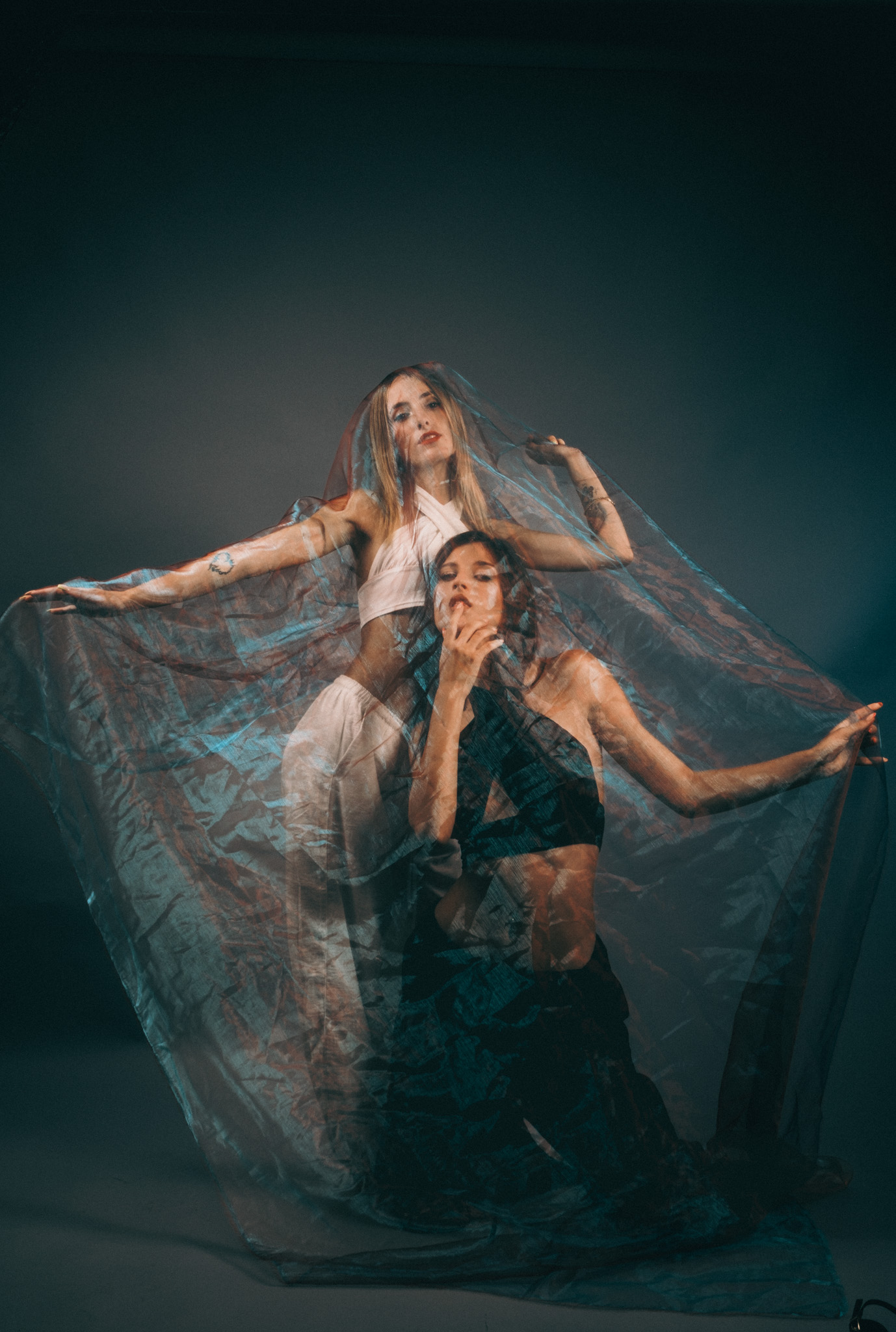 Two women in a studio holding a large sheer fabric in warm amber and teal tones, one kneeling while the other spreads it overhead