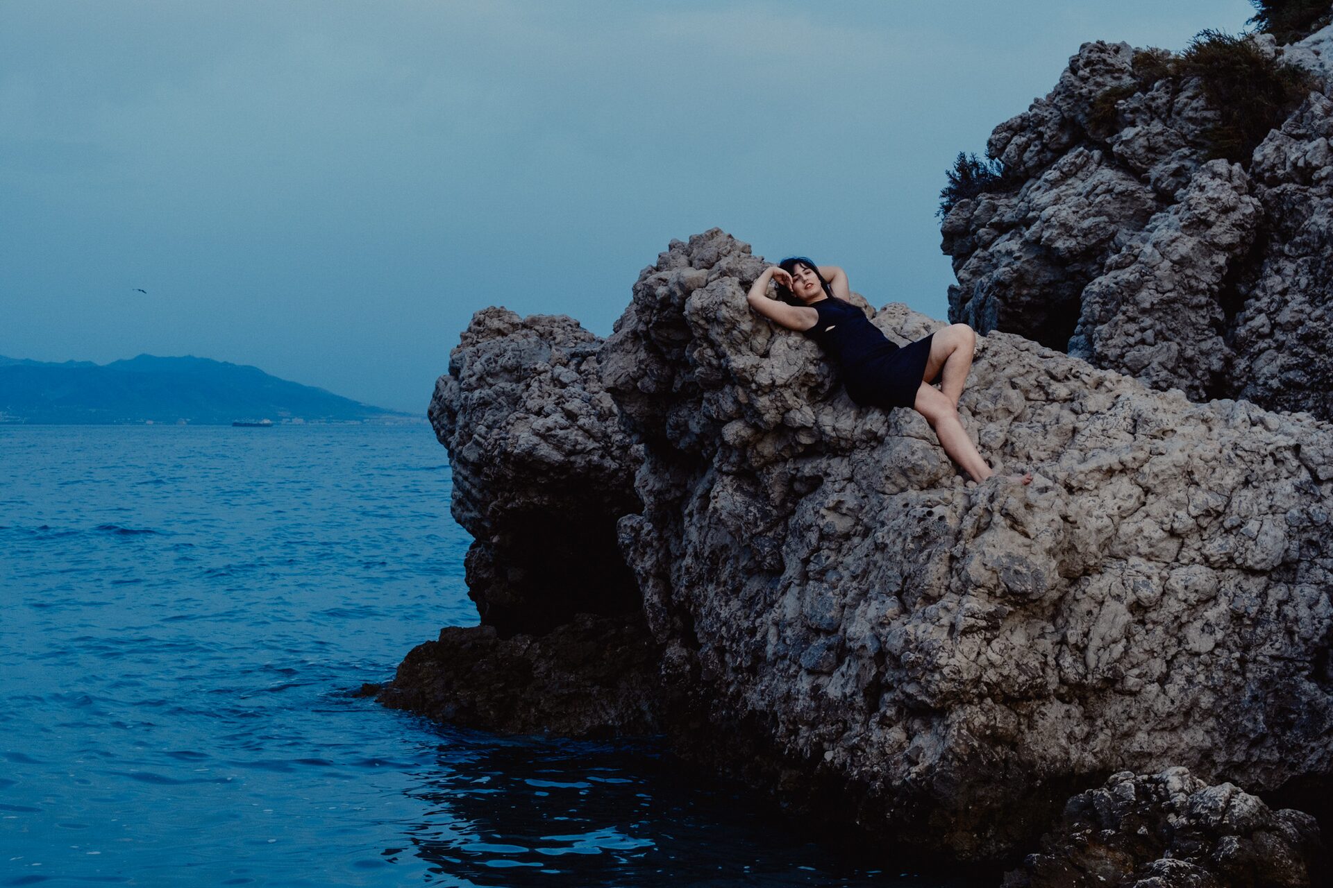 Woman reclining on rugged coastal rocks at dusk in a dark swimsuit, calm Mediterranean sea and distant hills behind her in the evening blue light
