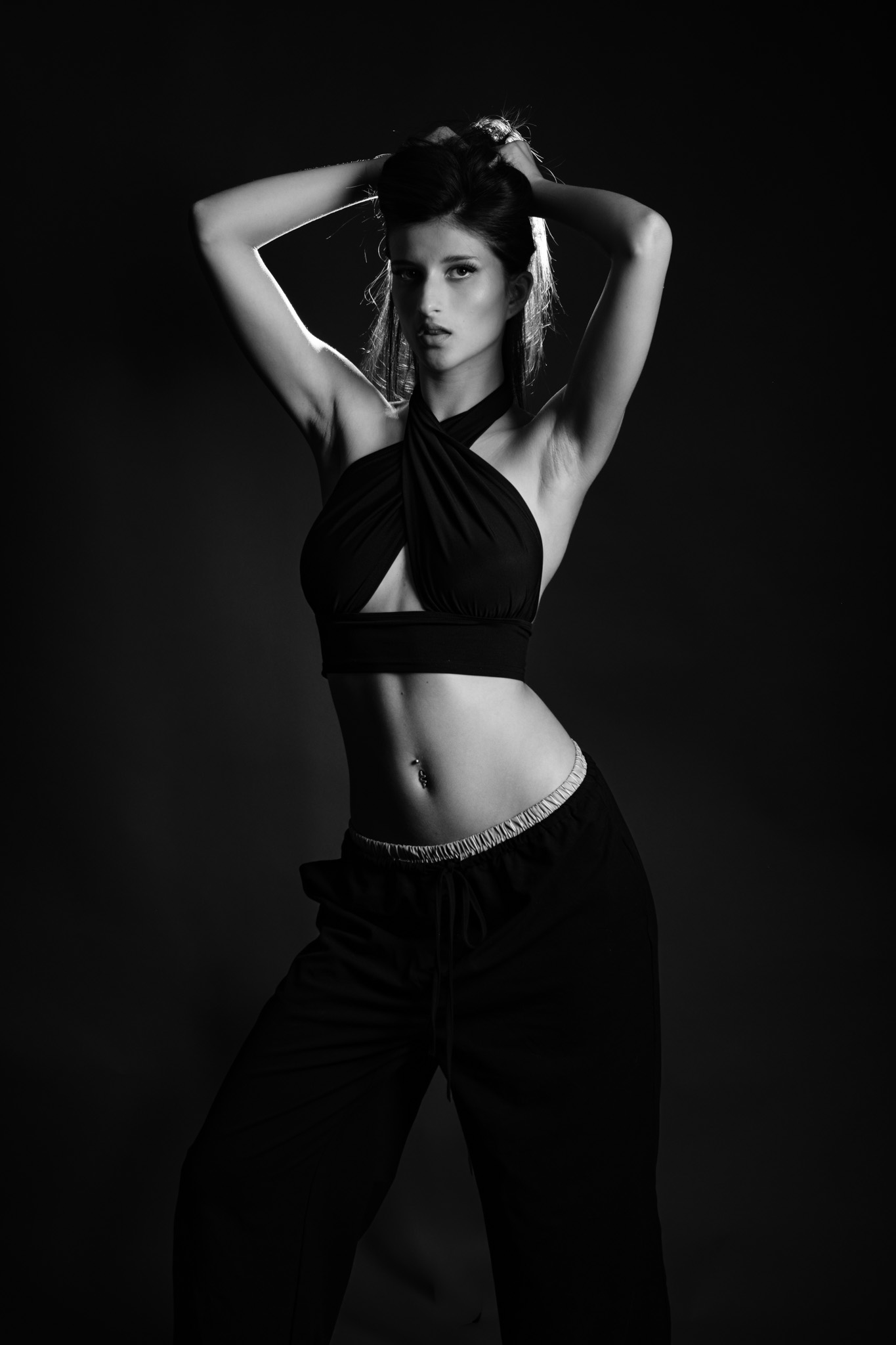 Studio portrait of a woman in a black cross-neck halter top, arms raised above her head, dark background, black and white